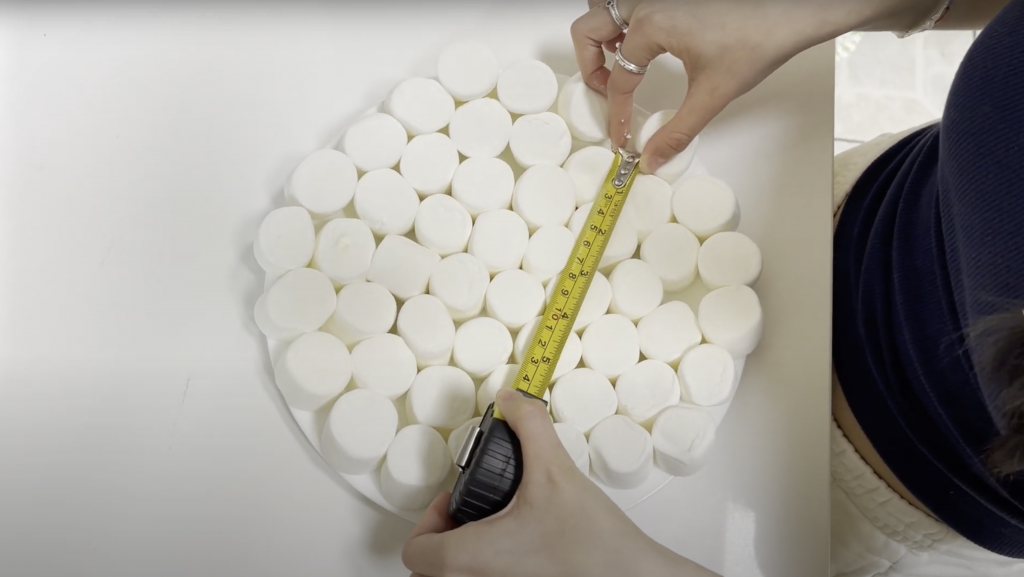 Close up, from above, of hands measuring (with a measuring tape) a plate of marshmallows on a white counter.