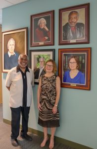 Two people standing in front of a wall with 5 portraits of former Directors displayed.